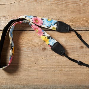 Op de afbeelding: Een camerariem met een bloemenpatroon in roze, geel en blauw op een witte achtergrond. De riem heeft een zwarte achterkant en verstelbare zwarte bandjes. De riem is ontworpen om aan een camera te worden bevestigd.