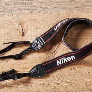 Può includere: Cinturino nero per fotocamera Nikon con dettagli bianchi e rossi. Il cinturino presenta il logo Nikon in bianco. Include cinturini neri regolabili per il fissaggio a una fotocamera. Realizzato in materiale resistente.
