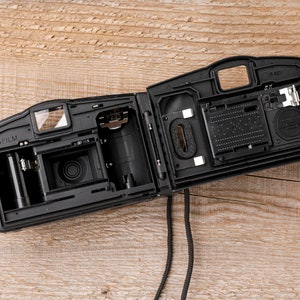 May include: Black, open film camera with internal components visible. The camera is on a wooden surface. The camera's interior includes a film compartment, lens, and other mechanical parts. A black strap hangs down.