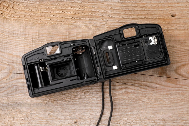 May include: Black, open film camera with internal components visible. The camera is on a wooden surface. The camera's interior includes a film compartment, lens, and other mechanical parts. A black strap hangs down.