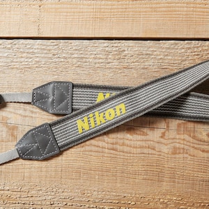 May include: A grey and white striped Nikon camera strap with yellow lettering. The strap has grey leather accents and black plastic buckles. The strap is lying on a wooden surface.