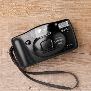 May include: Black Fuji DL-95 Super camera with a built-in flash and a wrist strap. The camera has the text "AUTO FOCUS DL-95 SUPER" and "MACRO" printed on it. The camera is on a wooden surface.