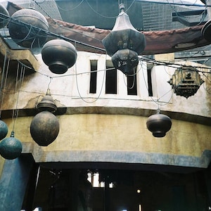 May include: An interior shot of a building with hanging spherical and lantern-shaped light fixtures. The lights are suspended from wires and cables. The walls are a mix of beige and gray, with a weathered appearance. The overall theme is rustic and industrial.