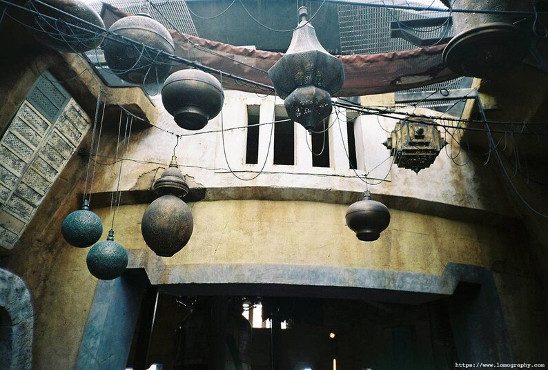 May include: An interior shot of a building with hanging spherical and lantern-shaped light fixtures. The lights are suspended from wires and cables. The walls are a mix of beige and gray, with a weathered appearance. The overall theme is rustic and industrial.
