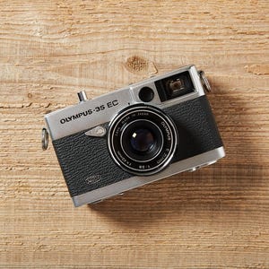 Può includere: Una fotocamera Olympus-35 EC con corpo argentato e impugnatura in pelle nera è appoggiata su una superficie di legno. L'obiettivo della fotocamera è centrato, con il nome del marchio stampato sopra. È un oggetto fotografico vintage.