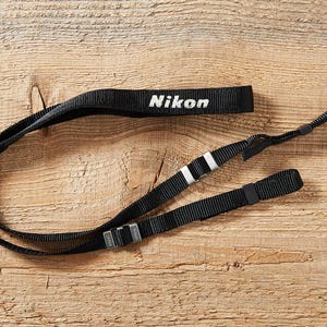 May include: A black camera strap made of woven nylon webbing featuring the white text Nikon printed on the main section. The strap includes adjustable metal buckles and black plastic sliders to modify the length for secure camera attachment.