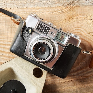 May include: A silver Agfa Color-Agnar camera with a 1:2.8/45 lens, encased in a black leather half-case with a brown leather neck strap. The camera features a textured black grip area, a viewfinder window, and a metal top plate with dials.