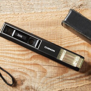 May include: A black Chinon pocket camera with a rectangular body, a built-in flash, and a lens labeled tele. The camera features a black wrist strap attached to one side and is accompanied by a matching black leather-like protective case.