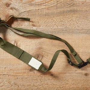 May include: Olive green camera strap with a white label and black plastic clips. The strap is made of a durable, woven material and is designed to be adjustable. The strap is lying on a wooden surface.