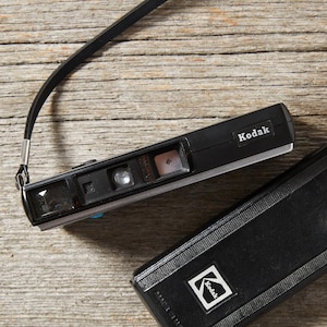 May include: A black Kodak film camera with a rectangular body, a lens, a viewfinder, and a black wrist strap. Next to it is a matching black rectangular camera case featuring a white Kodak logo on the front surface of the cover.