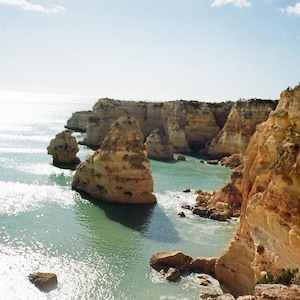 May include: Coastal landscape featuring a turquoise sea and towering, weathered rock formations. The image captures a sunny day with a clear blue sky, highlighting the natural beauty of the cliffs and water. The scene evokes a sense of tranquility and the power of nature.