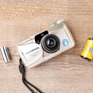 May include: A champagne-colored Olympus Stylus Epic Zoom 80 camera with a black wrist strap, a silver battery, and a yellow roll of Funtime Film 200. The camera features a lens, a flash, and a blue water-resistant icon on the front casing.
