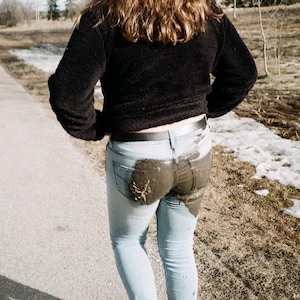May include: A person wearing a black sweater and light blue jeans with a large stain on the rear. The person is walking on a paved path next to a snowy area with trees in the background.