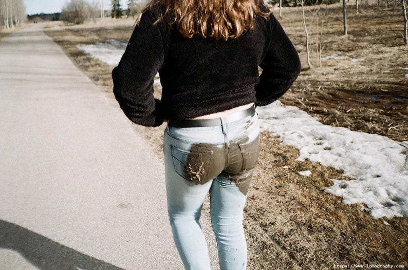 May include: A person wearing a black sweater and light blue jeans with a large stain on the rear. The person is walking on a paved path next to a snowy area with trees in the background.