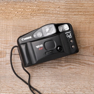 May include: Black Canon Sure Shot Owl Date 35mm film camera with a built-in flash and a black strap. The camera has the Canon logo and the text "Canon Lens 38mm 1:2.8 AF". The camera is on a wooden surface.