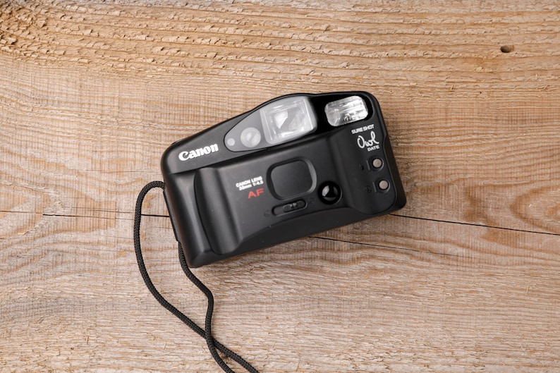 May include: Black Canon Sure Shot Owl Date 35mm film camera with a built-in flash and a black strap. The camera has the Canon logo and the text "Canon Lens 38mm 1:2.8 AF". The camera is on a wooden surface.