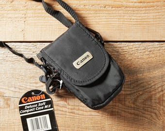 May include: A black Canon Deluxe Soft Compact Case M II camera bag with a flap closure, a metal logo plate, and an adjustable black strap. A black product tag is attached, featuring the Canon logo and the text Deluxe Soft Compact Case M II.