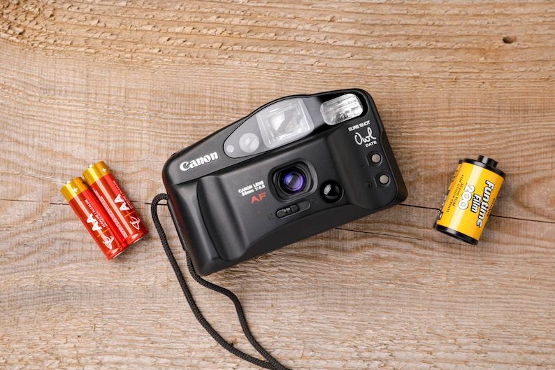 May include: A black Canon Sure Shot Owl Date 35mm AF camera with a lens, flash, and strap. Two red and gold AA batteries and a roll of Funtime 200 film are next to the camera. The items are on a wooden surface.