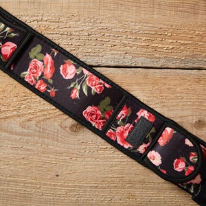 Op de afbeelding: Zwarte gitaarband met een bloemenrozenpatroon in rood en roze. De band heeft zwarte leren uiteinden en een gewatteerd gedeelte. Het bloemendesign staat tegen een zwarte achtergrond.