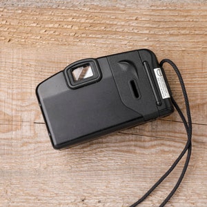 May include: Black point-and-shoot film camera with a viewfinder, flash, and a wrist strap. The camera is rectangular with rounded edges and a small label on the side. The camera is on a light brown wooden surface.