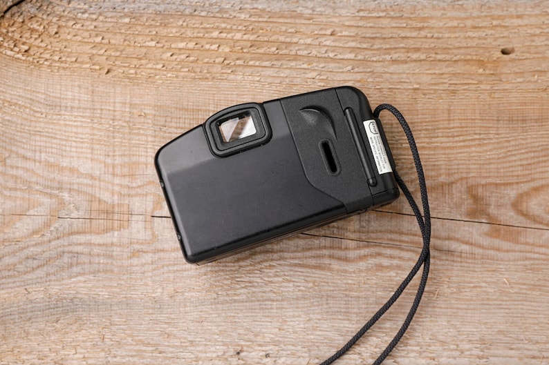 May include: Black point-and-shoot film camera with a viewfinder, flash, and a wrist strap. The camera is rectangular with rounded edges and a small label on the side. The camera is on a light brown wooden surface.