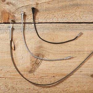 May include: Three camera shutter release cables, each with a different design, are arranged on a wooden surface. One is black, one is silver, and the other is black. The cables have a metal tip and a button.