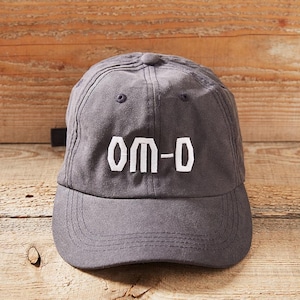 Może przedstawiać: Ciemnoszara czapka z daszkiem z haftowanym białym logo z napisem "OM-D".