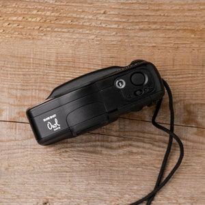 May include: A black Sure Shot Owl Date camera with a black strap lies on a wooden surface. The camera has a lens, buttons, and the text "Sure Shot Owl Date" in white. The camera is a vintage style.