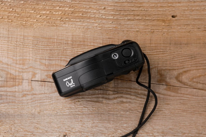 May include: A black Sure Shot Owl Date camera with a black strap lies on a wooden surface. The camera has a lens, buttons, and the text "Sure Shot Owl Date" in white. The camera is a vintage style.
