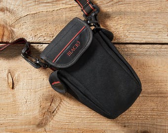 May include: Black camera bag with a red and black strap. The bag features a flap with the word "BLACKS" in red lettering. The bag is made of a textured material and has a small side pocket. It rests on a wooden surface.