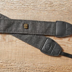 Op de afbeelding: Een grijze visgraat camerariem met zwarte leren uiteinden en een messingkleurige klinknagel. De riem heeft een zwarte nylon connector. De leren uiteinden hebben een ingeprent camera-icoon. De riem ligt op een houten oppervlak.