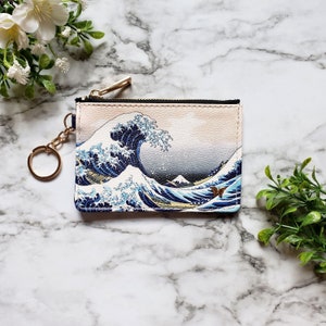 May include: A small pink and white zippered pouch with a gold chain keychain. The pouch features a printed image of the Great Wave off Kanagawa, a famous Japanese woodblock print by Hokusai.