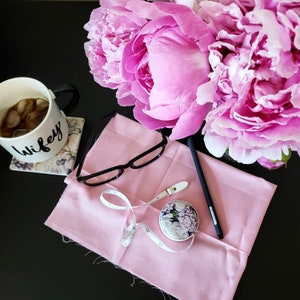 May include: A still life arrangement featuring a pink fabric square, a floral-patterned tape measure, black-framed glasses, and a black pen. A mug with the word "Wifey" and a coaster are next to a bouquet of pink peonies. The items are on a black surface.
