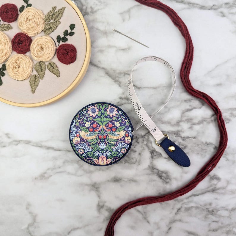 K&ouml;nnte beinhalten: Ein rundes, marineblaues Ma&szlig;band mit einem floralen und Vogelmuster. Ein wei&szlig;es Ma&szlig;band erstreckt sich aus dem Geh&auml;use. Ein Stickrahmen mit floraler Stickerei und eine Nadel sind ebenfalls zu sehen, zusammen mit bordeauxrotem Faden.