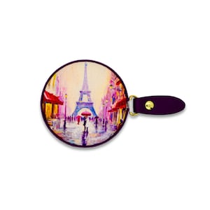 May include: A round, purple-bordered hand mirror featuring a painting of the Eiffel Tower in Paris. The artwork depicts a rainy street scene with people, red awnings, and colorful buildings. The mirror has a matching purple handle with a gold-colored clasp.