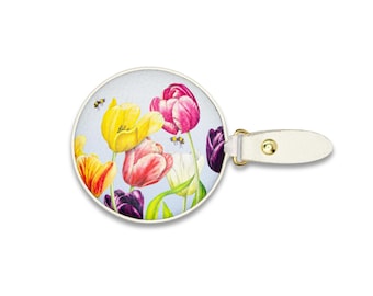 May include: A round luggage tag featuring a watercolor illustration of yellow, pink, white, and purple tulips with two small bees on a light blue background, attached to a white leather strap with a gold-toned metal snap button fastener.