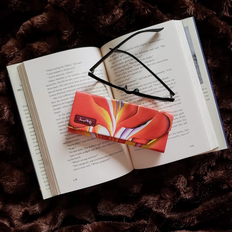 Pu&ograve; includere: Un libro aperto con occhiali con montatura nera e un astuccio per occhiali. L'astuccio presenta un colorato motivo floreale arancione, giallo e bianco. Il testo "Sweet Chef" &egrave; visibile sull'astuccio.