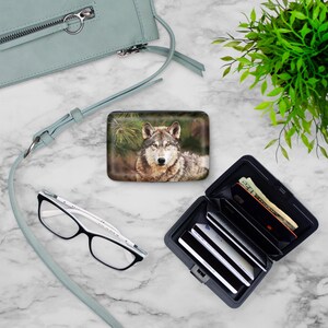 Peut inclure: Un portefeuille en m&eacute;tal noir avec un motif de loup sur le devant. Le portefeuille est ouvert et montre plusieurs cartes de cr&eacute;dit et de l'argent liquide &agrave; l'int&eacute;rieur. Le portefeuille est pos&eacute; sur une surface en marbre blanc.