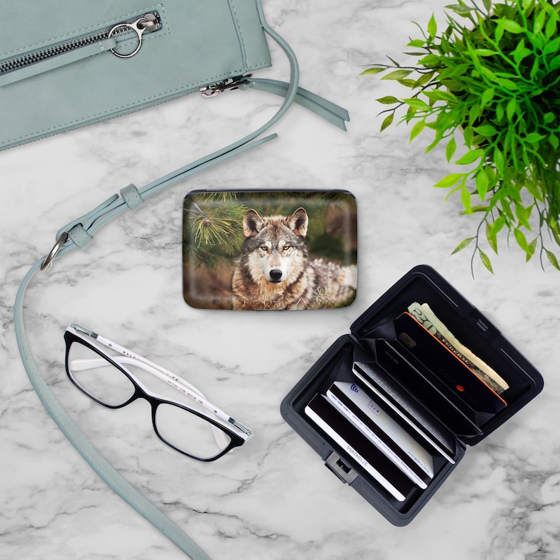 Peut inclure: Un portefeuille en m&eacute;tal noir avec un motif de loup sur le devant. Le portefeuille est ouvert et montre plusieurs cartes de cr&eacute;dit et de l'argent liquide &agrave; l'int&eacute;rieur. Le portefeuille est pos&eacute; sur une surface en marbre blanc.