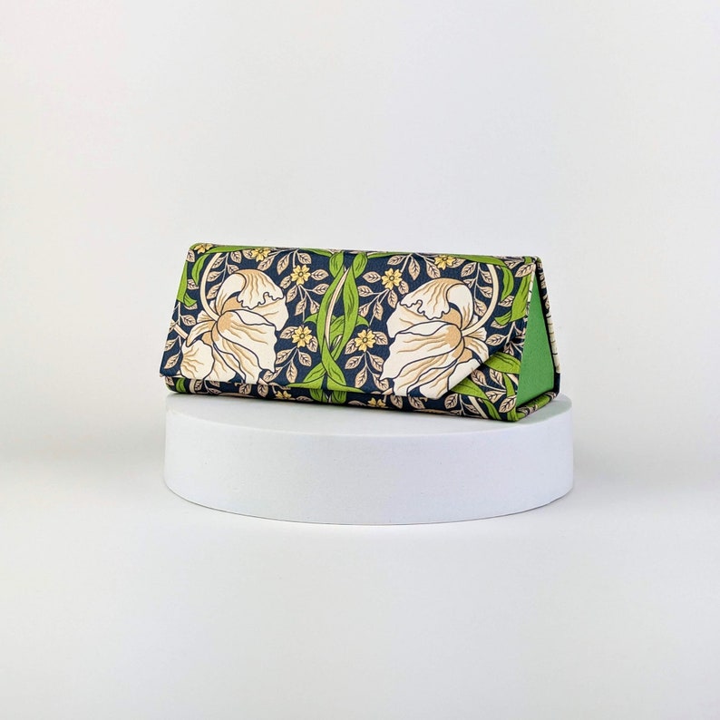 Op de afbeelding: Een driehoekig brillenetui met een bloemenpatroon. De etui heeft een marineblauwe achtergrond met cr&egrave;mekleurige bloemen, groene bladeren en kleine gele bloesems. De binnenkant is heldergroen.