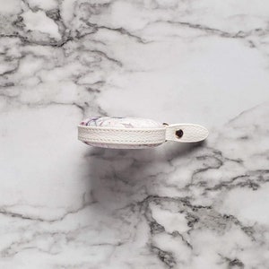 May include: A small, round, white leather case with a floral design in shades of purple and pink. The case has a white leather strap with a gold-colored snap closure. The edges are trimmed with white stitching.
