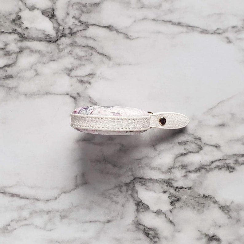 May include: A small, round, white leather case with a floral design in shades of purple and pink. The case has a white leather strap with a gold-colored snap closure. The edges are trimmed with white stitching.