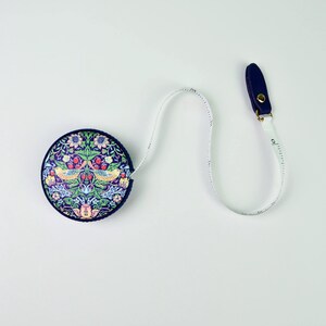 K&ouml;nnte beinhalten: Rundes, marineblaues Ma&szlig;band mit Blumen- und Vogelmuster. Das Band erstreckt sich von dem runden Geh&auml;use, mit einer marineblauen Kunstlederlasche am Ende. Das Band ist wei&szlig; mit schwarzen Messmarkierungen.