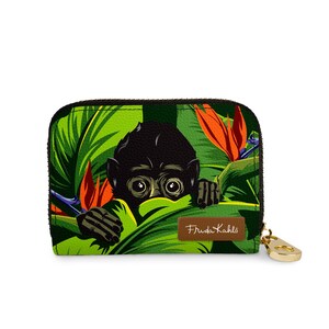 May include: A zippered wallet featuring a black monkey peeking through lush green leaves and orange flowers. The design includes a gold-colored zipper pull and a brown label with the text "Frida Kahlo".