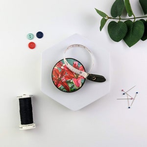 May include: A floral patterned tape measure with a retractable tape. The tape measure is round and has a black leather strap. The floral pattern features pink and red lilies on a blue background.
