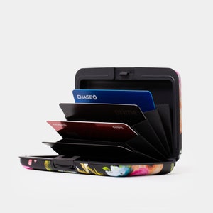 May include: A black metal card holder with a floral print design. The holder is open and shows multiple cards inside, including a Chase credit card, a Prime card, and a debit card.