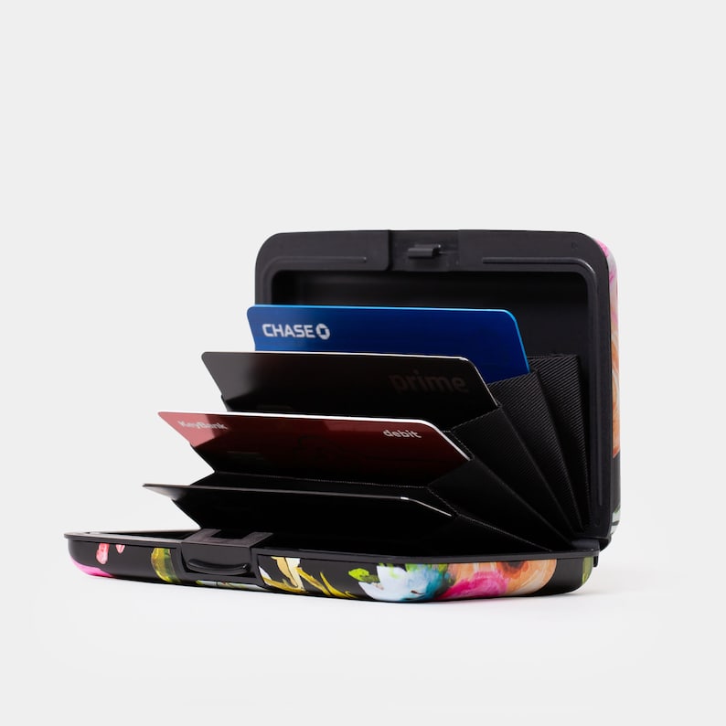 May include: A black metal card holder with a floral print design. The holder is open and shows multiple cards inside, including a Chase credit card, a Prime card, and a debit card.