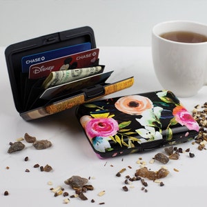 May include: Two metal card holders, one with a floral design and the other with a wood grain design. The floral design is black with pink, orange, and white flowers. The wood grain design is gold and brown. Both card holders are open and show the interior slots for cards.