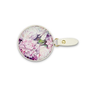 May include: Round compact mirror with a floral peony design in shades of pink, purple, and white. The mirror has a gold-colored rim and a white leatherette handle with a gold-colored rivet.