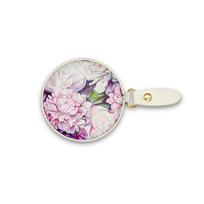 May include: Round compact mirror with a floral peony design in shades of pink, purple, and white. The mirror has a gold-colored rim and a white leatherette handle with a gold-colored rivet.